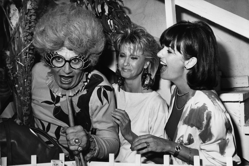 Dame Edna, Olivia Newton John and Pat Carroll stand together laughing in black and white. 