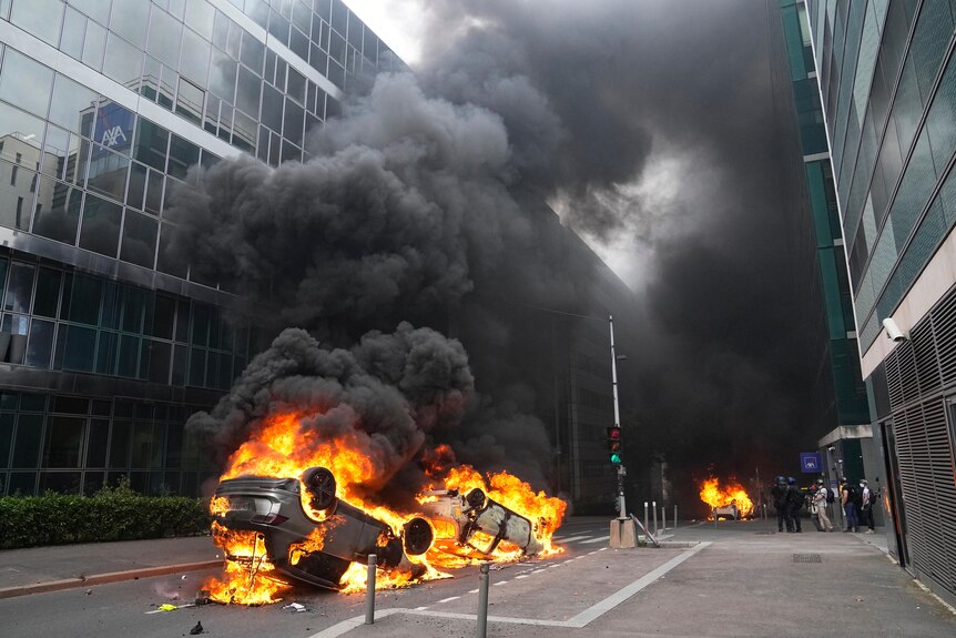 Burning cars are pictured upside down in the street while large plumes of smoke come off it.