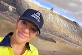 A close-up a smiling, 30-plus woman, in blue cap with Nara Nation logo in a blue and yellow top standing in front of a lake.