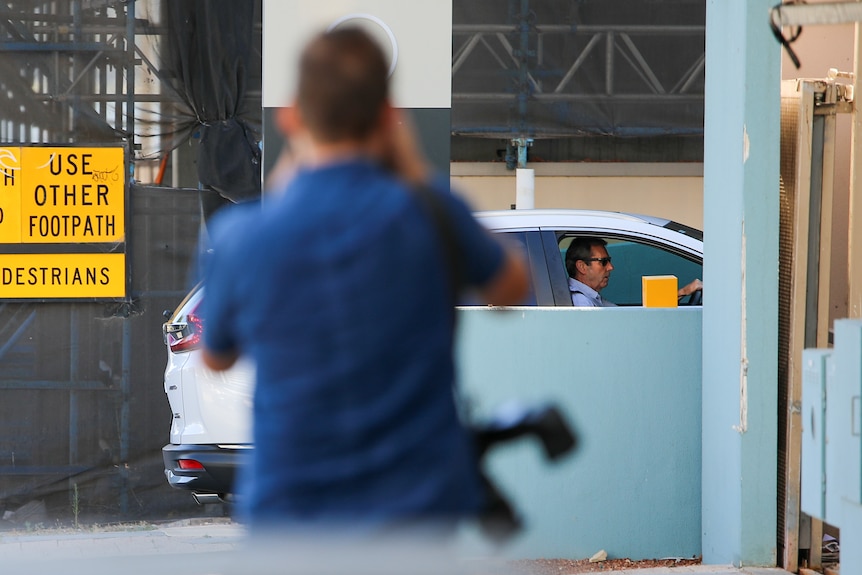 A photographer stands out of focus in the foreground taking a picture of a man driving a car.