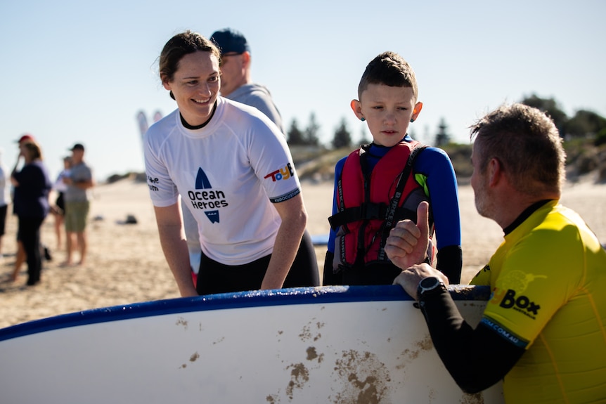 A young boy standing next to a surfboard looks at a man in a yellow rashie giving him instructions.