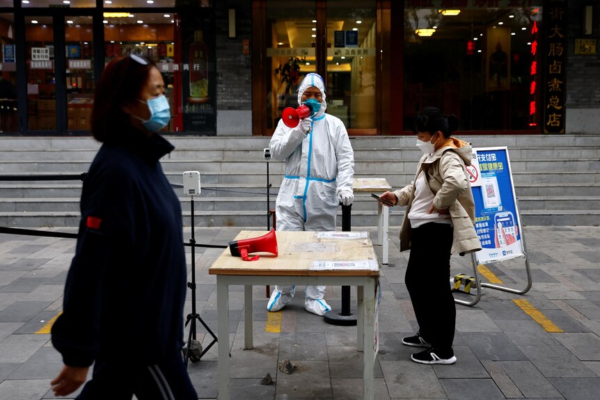 A worker in a protective suit guides people to scan a QR health code.