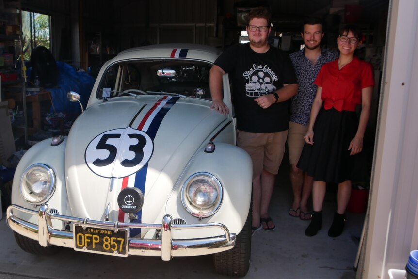 A cream vintage car with "53" on it, flanked by two mean and a woman.