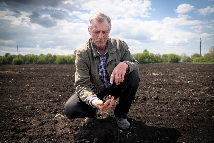 A man with grey hair and wearing a green jacket squats as he holds some soil in his hand.