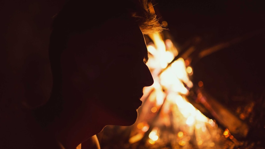 The silhouette of a mans face against an orange burning fire in the background.