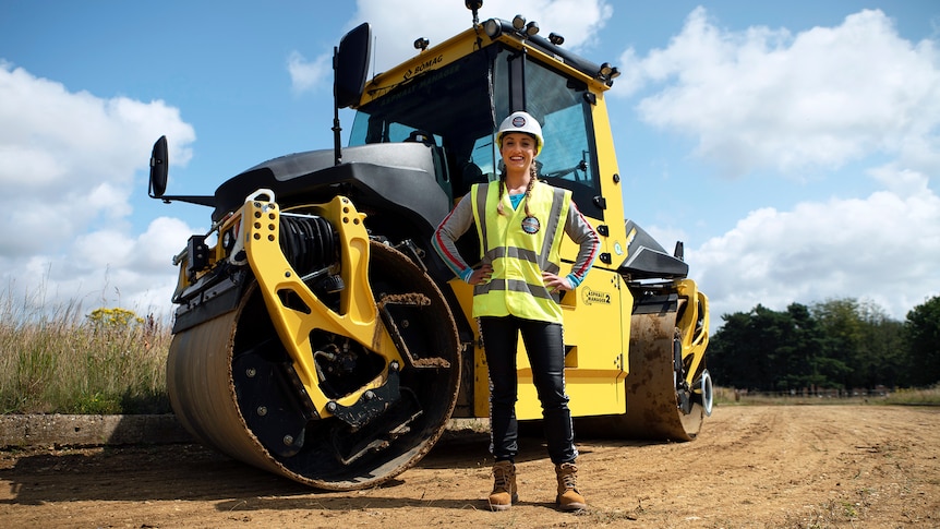 A woman in a hard hat stands in front of a large road-building machine