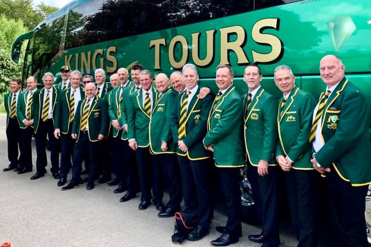 a line of men in their 60s wearing Green and gold jackets stand in front of a bus