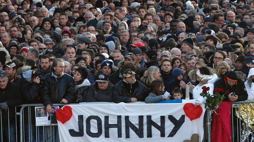 Hundreds of people crowd outside a fence bearing a sign which says Johnny with pictures of two hearts outside Hallyday's funeral