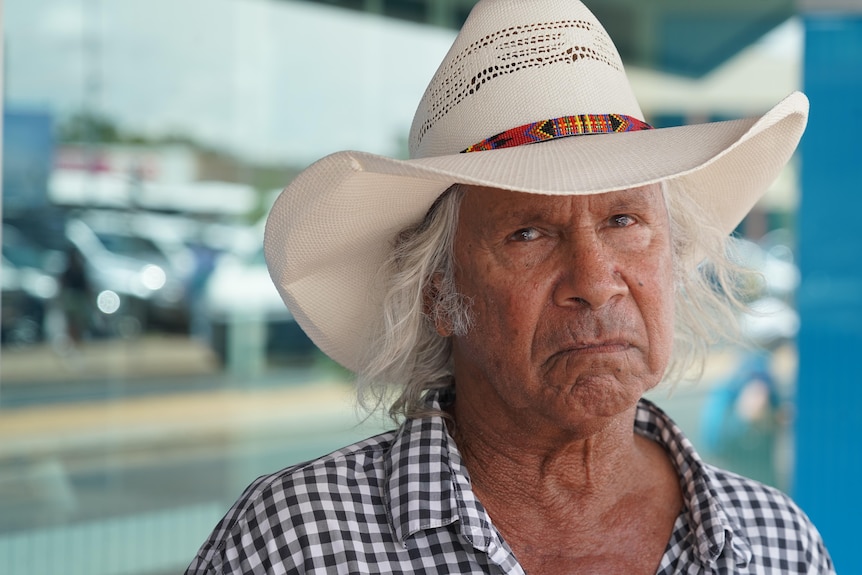 A man with a white hat looks seriously at the camera. He has white hair and a checked shirt.