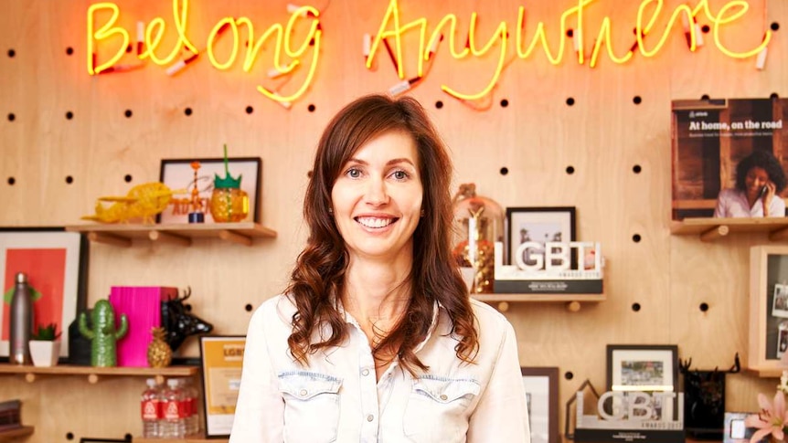 A woman with long brown hair smiles against a backdrop that reads belong anywhere in neon