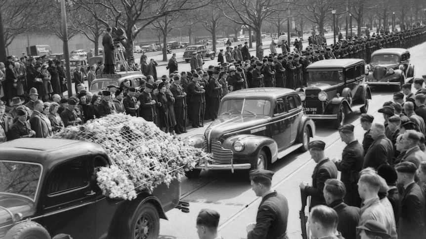 Black and white photo of crowds gathering around old cars carrying the coffins of the victims.