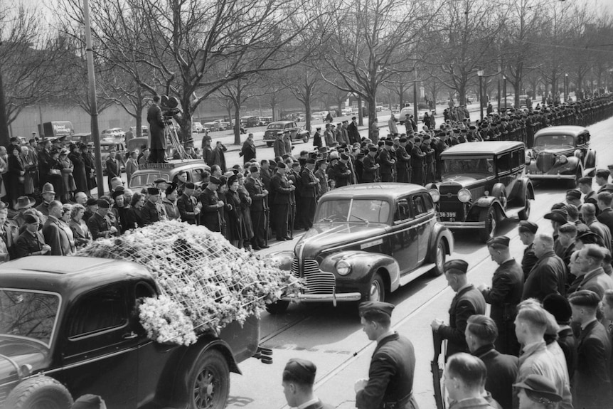 Black and white photo of crowds gathering around old cars carrying the coffins of the victims.