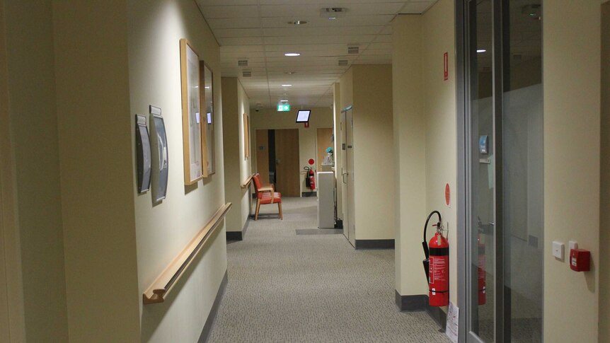 A hospital hallway with a fire extinguisher in the foreground and chairs and an exit sign in the distance.