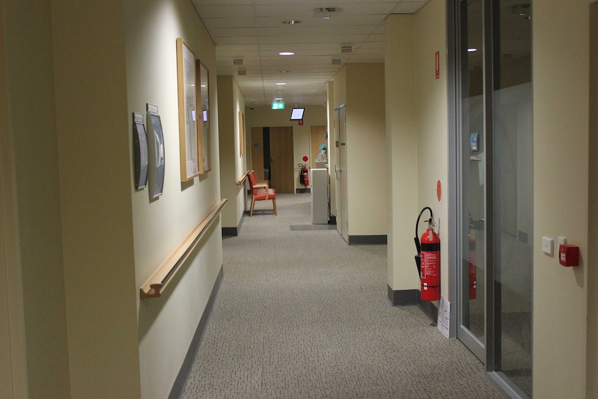 A hospital hallway with a fire extinguisher in the foreground and chairs and an exit sign in the distance.