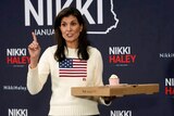 Nikki Haley wears a sweater with the american flag and holds a pizza box as she speaks, pointing her right index finger up