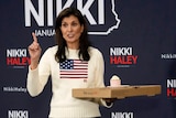 Nikki Haley wears a sweater with the american flag and holds a pizza box as she speaks, pointing her right index finger up