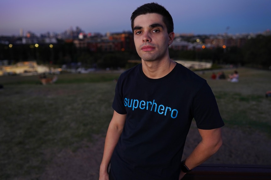 Man wearing a black shirt with the text 'superhero', standing in a park.