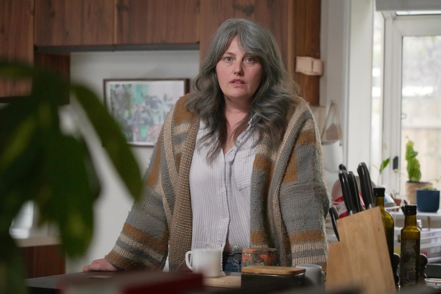 A white woman with greying brown hair standing in a kitchen with wooden cabinets