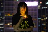 A woman wearing a black t-shirt and glasses stands in a high-rise building holding a battery-powered candle looking solemn