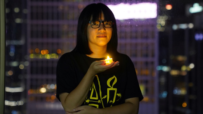 A woman wearing a black t-shirt and glasses stands in a high-rise building holding a battery-powered candle looking solemn