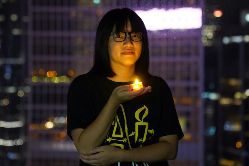 A woman wearing a black t-shirt and glasses stands in a high-rise building holding a battery-powered candle looking solemn