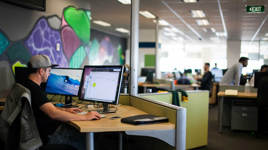 A man in a baseball hat sits at a desk next to a mural