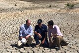 Three men crouch in the middle of a empty, cracked-earth dam
