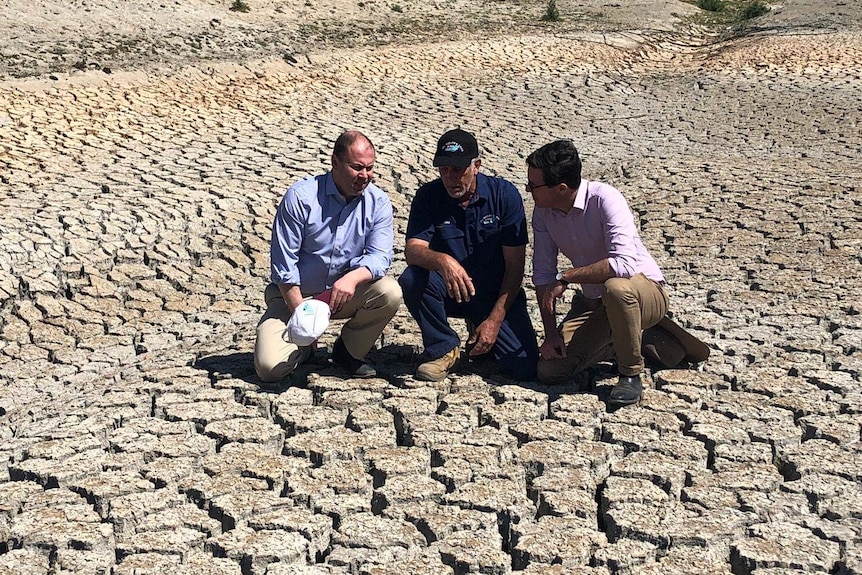 Three men crouch in the middle of a empty, cracked-earth dam.