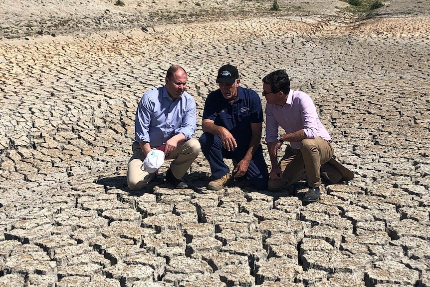 Three men crouch in the middle of a empty, cracked-earth dam