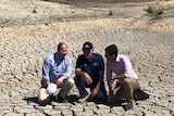 Three men crouch in the middle of a empty, cracked-earth dam.