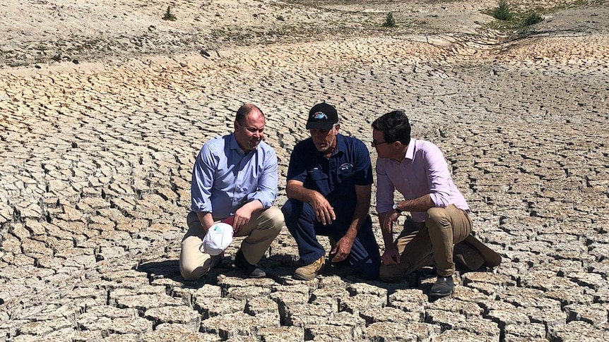 Three men crouch in the middle of a empty, cracked-earth dam