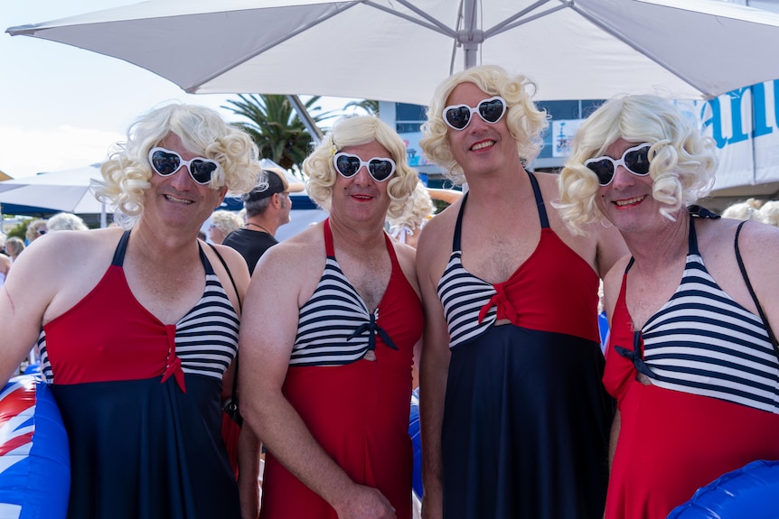 Four people wearing red and black swimwear, blonde curly wigs and white heart-shaped sunglasses stand under a white umbrella