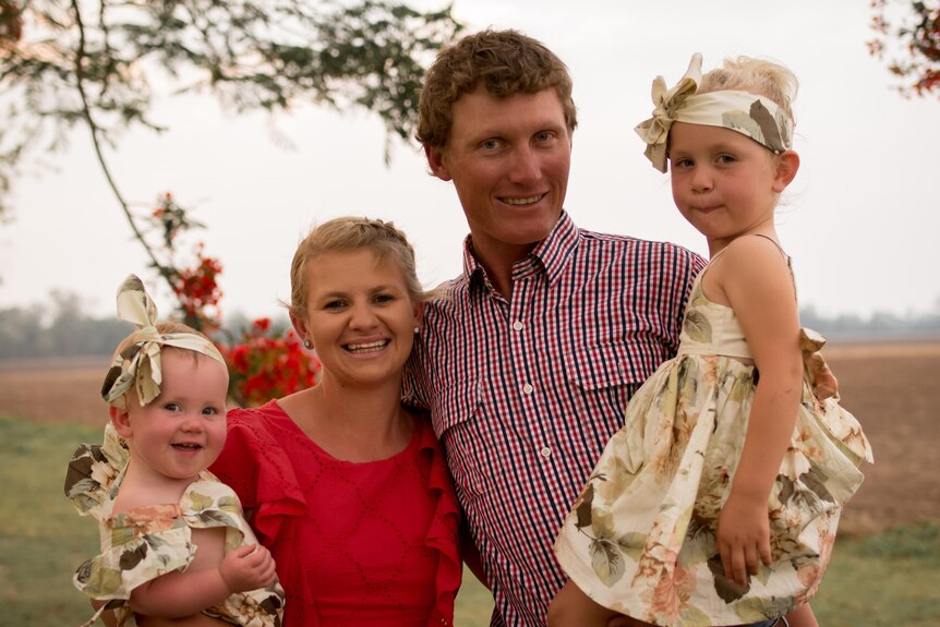 Kim Stevens stands next to her husband, both are holding their young daughters, with trees and fields visible in the background.