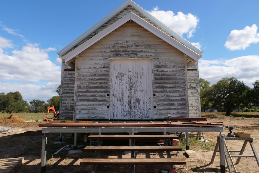 An old weathered wooden building with sloping roof, set on a raised platform with steps leading to it, under a blue sky.