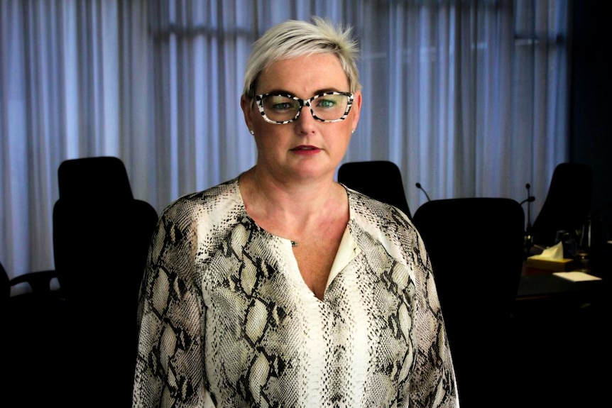 A woman with short hair and glasses stands in front of a desk fitted with microphones.