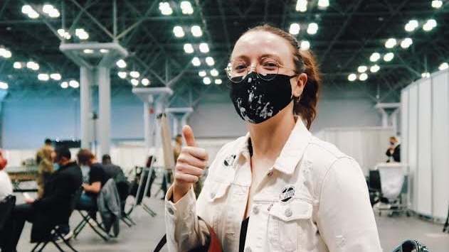 A woman wearing a cream jacket and face mask gives a thumbs up