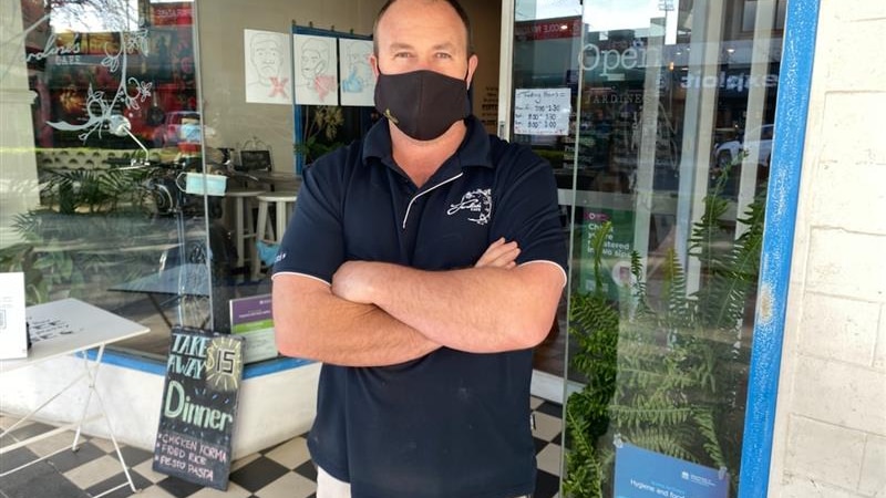 A cafe owner in a mask stands outside his business wearing a black mask