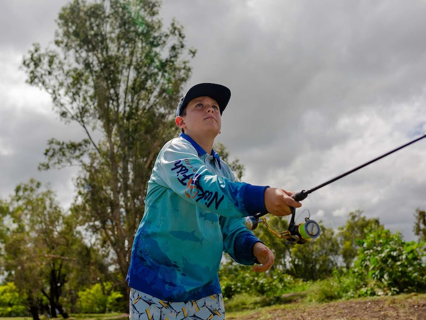 A young boy casts a line. Wearing blue cap and shirt, shark shorts. Grey clouds in background.