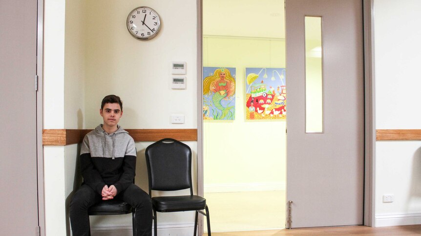 Matthew sits on a chair in a community hall below a clock hanging on the wall.