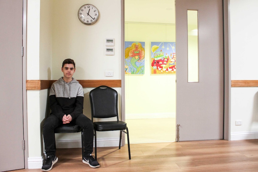 Matthew sits on a chair in a community hall below a clock hanging on the wall.