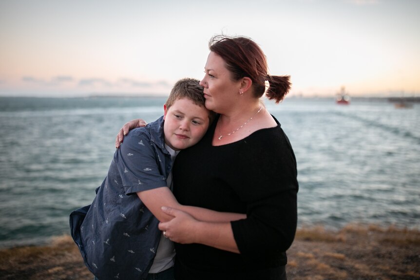 A boy rests his head on his mum as he puts his arms around her. The ocean is behind them.