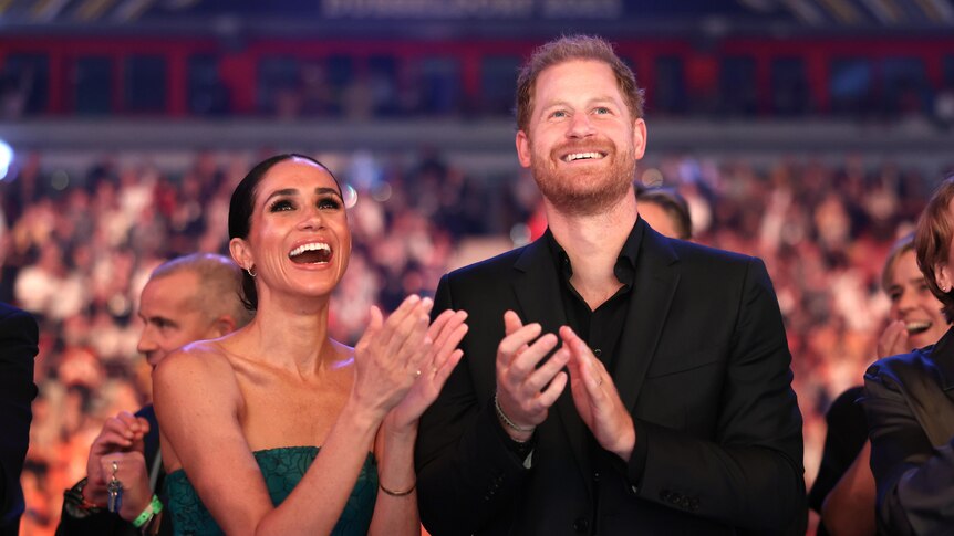Prince Harry and Meghan smiling at a function standing up and clapping