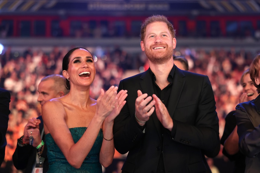 Prince Harry and Meghan smiling at a function standing up and clapping