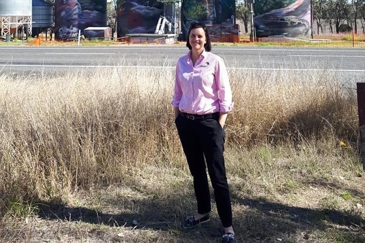 A dark haired smiling woman stands in front of painted silos She's wearing a pink pin striped shirt tucked into black pants