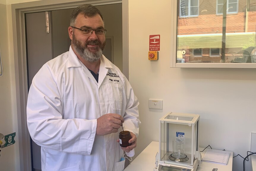 A man in a lab coat stands with a pot of honey.