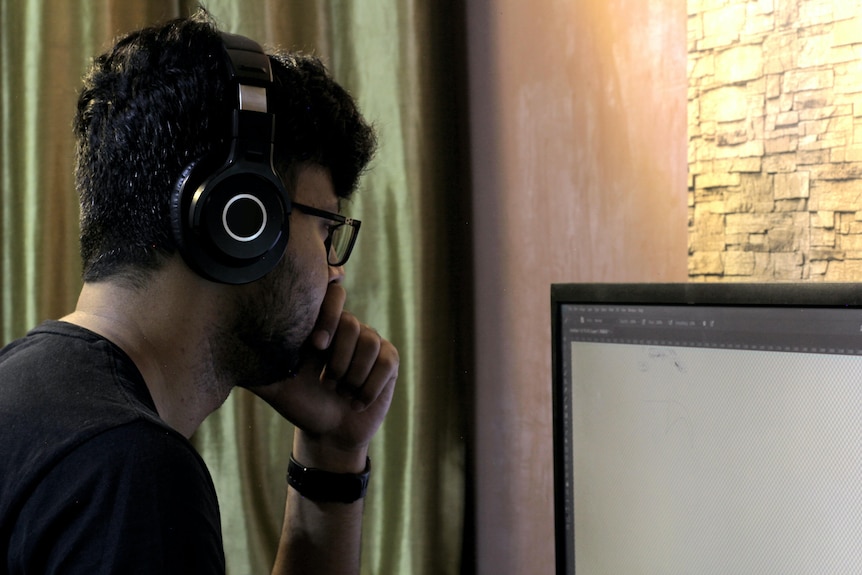 The side of a man's face in a darkened room. He wears headphones and looks at a computer screen.