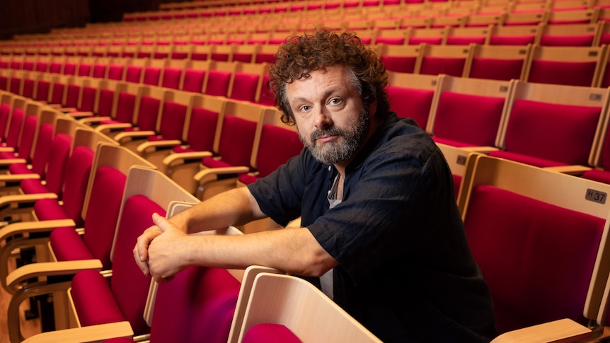 Michael Sheen, sitting in the middle of an empty auditorium, looks pensively at the camera.