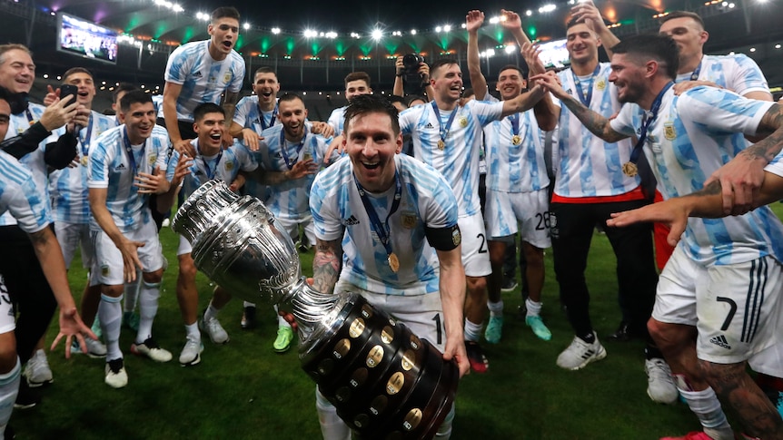 A grinning Lionel Messi leans forward into the camera, holding the Copa America trophy that his Argentina team has won.