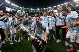 A grinning Lionel Messi leans forward into the camera, holding the Copa America trophy that his Argentina team has won.