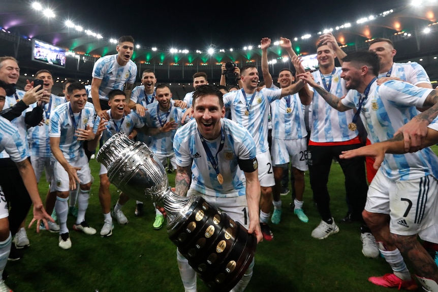 A smiling Lionel Messi leans forward towards the camera, holding the Copa America trophy his Argentina team have won.